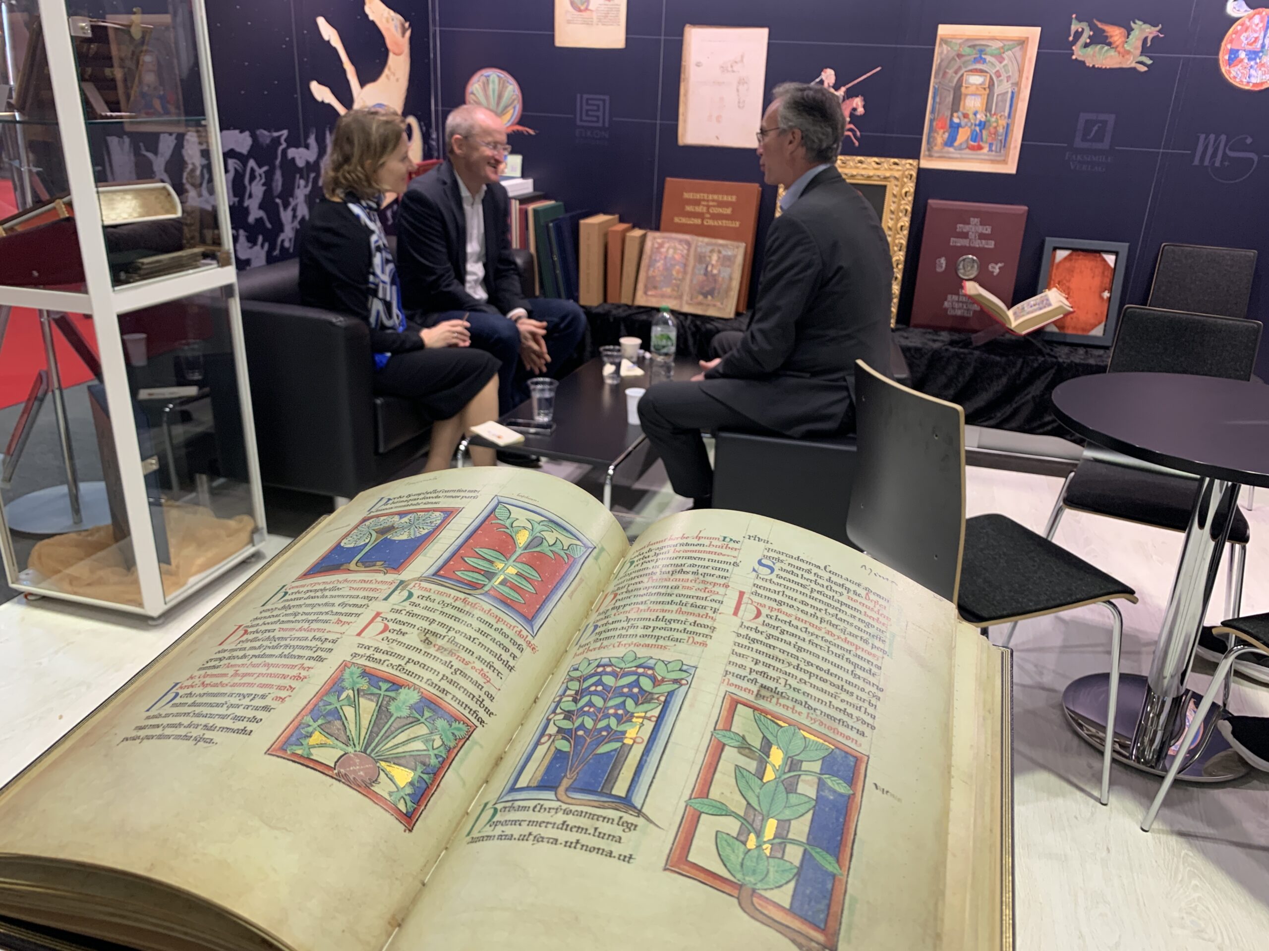 Frankfurter Buchmesse 2023: Im Vordergrund das aufgeschlagene Faksimile "Von Pflanzen und Tieren" mit fünf wundervollen Miniaturen von verschiedenen Heilpflanzen. Im Hintergrund unsere Verlegerin vertieft im Gespräch mit zwei Personen.